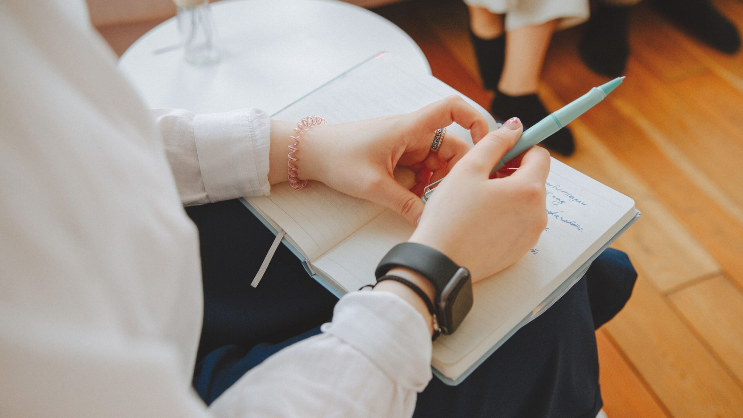 A person wearing a smartwatch writes in a notebook with a light blue pen.