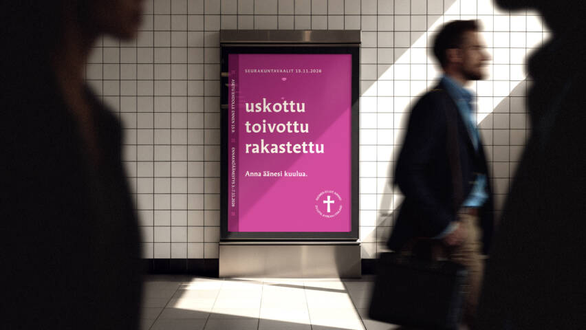 Kaupunkitilan seinällä oleva digitaalinen mainostaulu, jossa violetti juliste. Julisteessa teksti: ‘uskottu, toivottu, rakastettu – anna äänesi kuulua’. Etualalla ohikulkijoita liikkeessä.