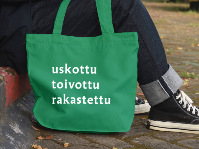 Vihreä kangaskassi, jossa teksti ‘uskottu, toivottu, rakastettu’. Kassi nojaa katukiveykseen istuvan henkilön vieressä.