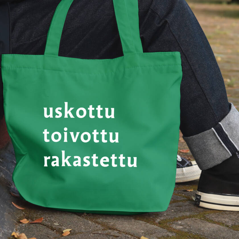 Vihreä kangaskassi, jossa teksti ‘uskottu, toivottu, rakastettu’. Kassi nojaa katukiveykseen istuvan henkilön vieressä.