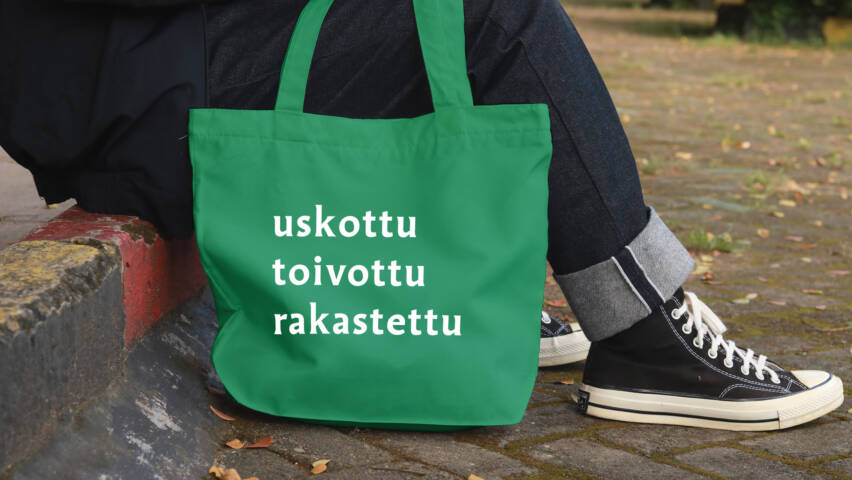Vihreä kangaskassi, jossa teksti ‘uskottu, toivottu, rakastettu’. Kassi nojaa katukiveykseen istuvan henkilön vieressä.