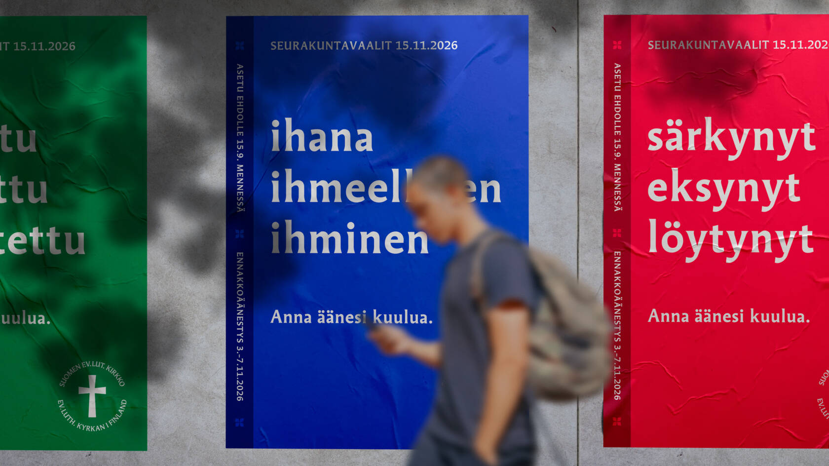 Kaupunkiseinällä kolme värikästä julistetta seurakuntavaaleista. Keskimmäisessä sinisessä julisteessa teksti: ‘ihana, ihmeellinen ihminen – anna äänesi kuulua’. Etualalla ohikulkija kävelee julisteiden ohi.