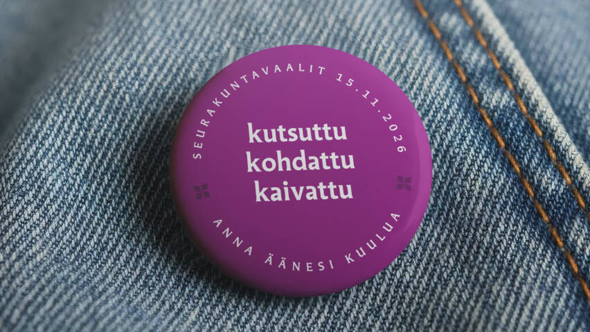 Violetti rintamerkki farkkukankaalla. Rintamerkissä teksti: ‘Seurakuntavaalit 15.11.2026 – kutsuttu, kohdattu, kaivattu – anna äänesi kuulua’