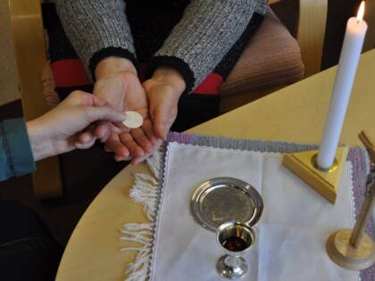Kädet vastaanottavat ehtoollisleipää toisen henkilön käsistä; pöydällä näkyvät ehtoolliskalkki punaisella nesteellä, hopeinen lautanen, palava kynttilä ja puinen krusifiksi.