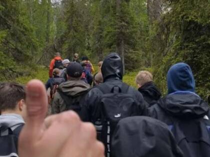 Ryhmä ihmisiä kävelee metsäpolkua pitkin. Etualalla näkyy peukku pystyssä -ele.