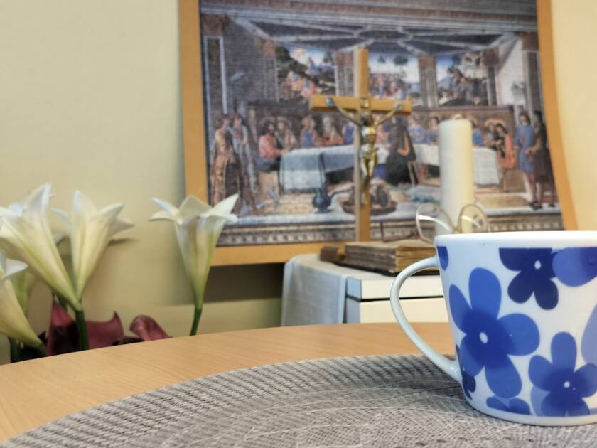 Sinivalkoinen kukkakuvioinen muki on pöydällä, taustalla kehystetty taulu, risti ja valkoisia kukkia.