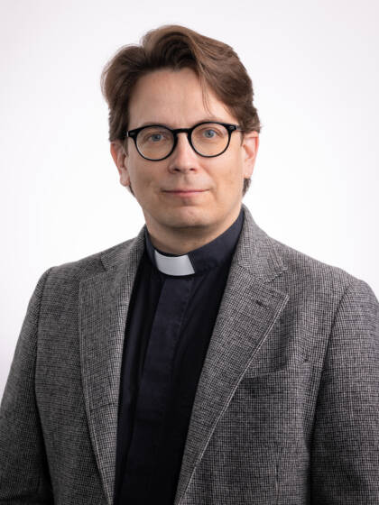 Silmälasipäinen miespappi, jolla harmaa takki mustan papinpaidan päällä.
