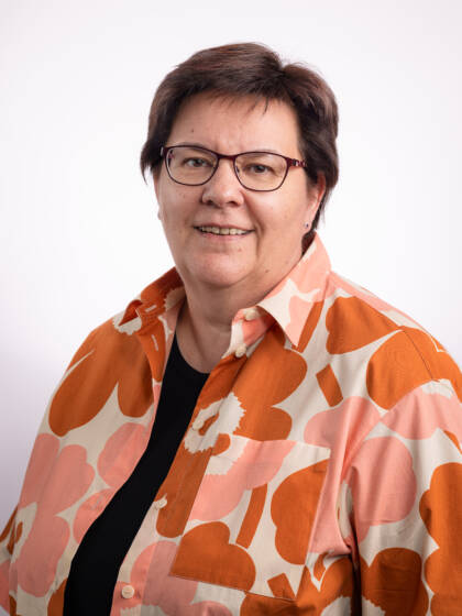 Hymyilevä silmälasipäinen nainen, jolla lyhyet ruskeat hiukset ja kauluspaita, jossa oransseja suuria kukkia.