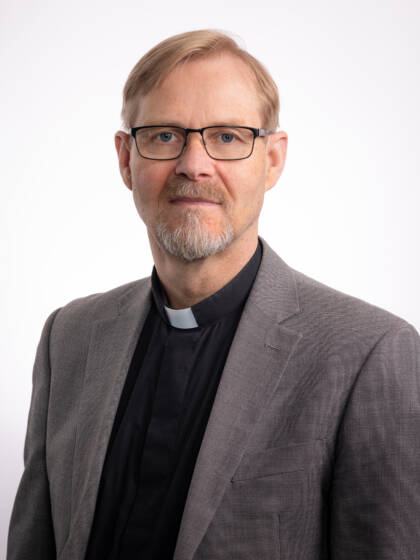 Vaaleahiuksinen silmälaisipäinen miespappi, jolla harmaa takki mustan papinpaidan päällä.