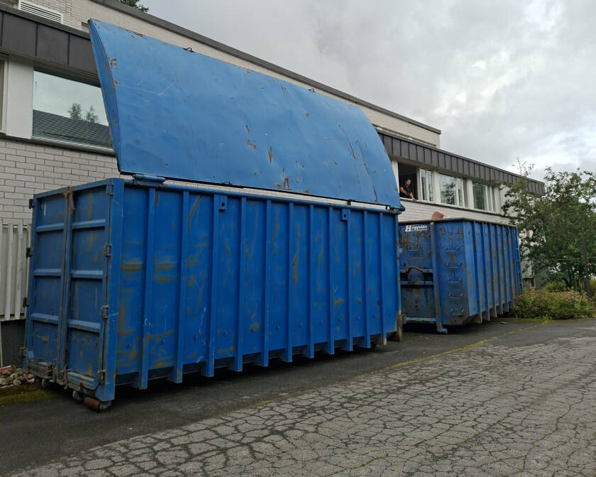 Kaksi sinistä roskalavaa rakennuksen edessä. Toiseen mies heittää tavaraa rakennuksen ikkunasta.