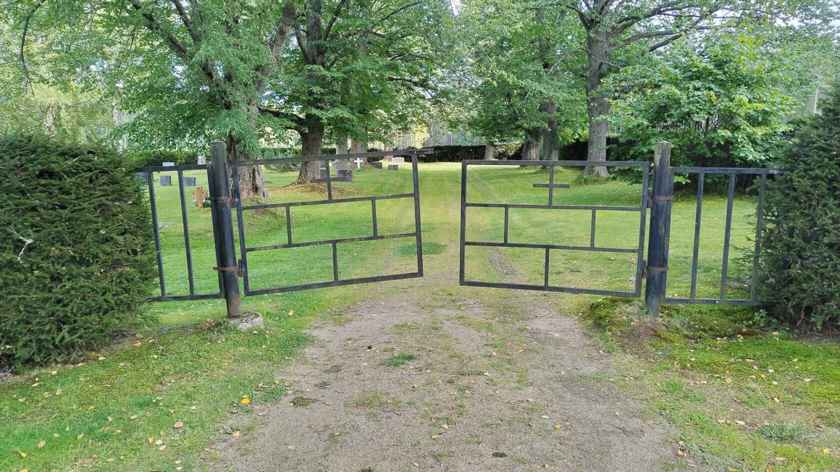 Kuhan hautausmaan sisäänkäyntiportti. Portin takana näkyy hautakiviä, nurmikkoa ja suuria puita. Hautausmaalle johtaa hiekkatie.