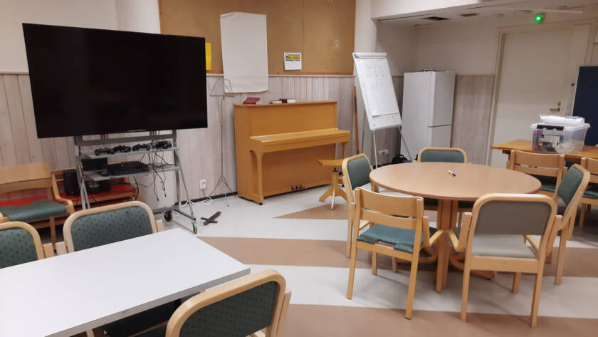 Nuorisotilassa televisio, piano ja pöytiä, joiden ympärillä tuoleja